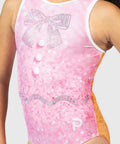 A close-up of a pink gymnastics leotard with a sparkling bow design on the chest, white buttons, a wavy rhinestone trim, and orange flip-side panel, shown on a model.