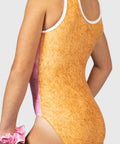 Close-up back view of a model wearing a one-piece swimsuit with a textured orange-brown body, white trim, and pink side panels, against a light gray background.