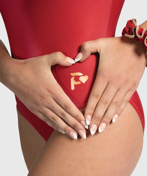 A close-up of a model wearing a red gymnastics leotard, with both hands forming a heart shape over the front. The center of the heart is printed with a gold logo featuring a stylized letter 'F' and a heart.