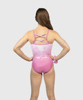 A girl from behind wearing a pink one-piece swimsuit with a glossy finish, floral pattern on the upper back, and crisscross straps, set against a solid light gray background.