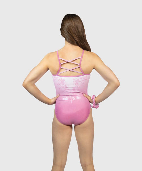 A girl from behind wearing a pink one-piece swimsuit with a glossy finish, floral pattern on the upper back, and crisscross straps, set against a solid light gray background.
