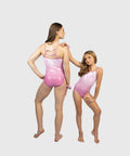 An adult woman and a young girl modeling shiny light pink leotards against a plain light gray studio background. The woman is shown from the back, wearing a strappy leotard, while the girl stands beside her in a sleeveless one-piece leotard.