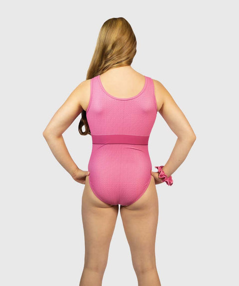 A girl with long blonde hair is shown from the back wearing a pink sleeveless one-piece leotard with a textured pattern and a solid pink waistband, set against a solid light gray background.